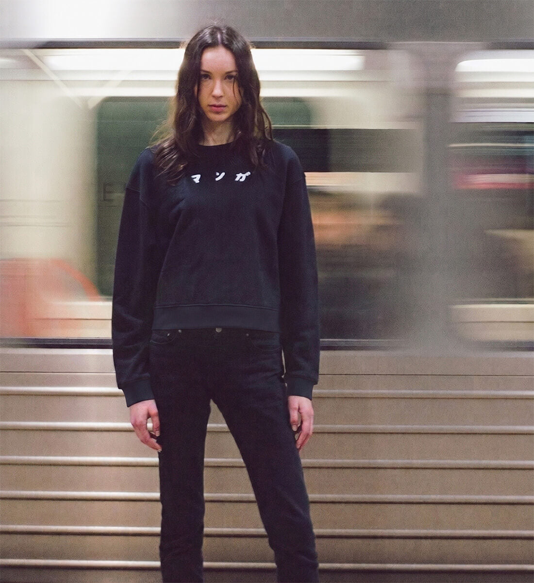 Urbane Szene an einem Bahnhof: Eine junge Frau steht vor einem vorbeifahrenden, unscharfen Zug. Sie trägt ein schwarzes, locker geschnittenes Boxy Sweatshirt mit einem weißen, japanischen Katakana-Schriftzug für Manga auf der Brust.