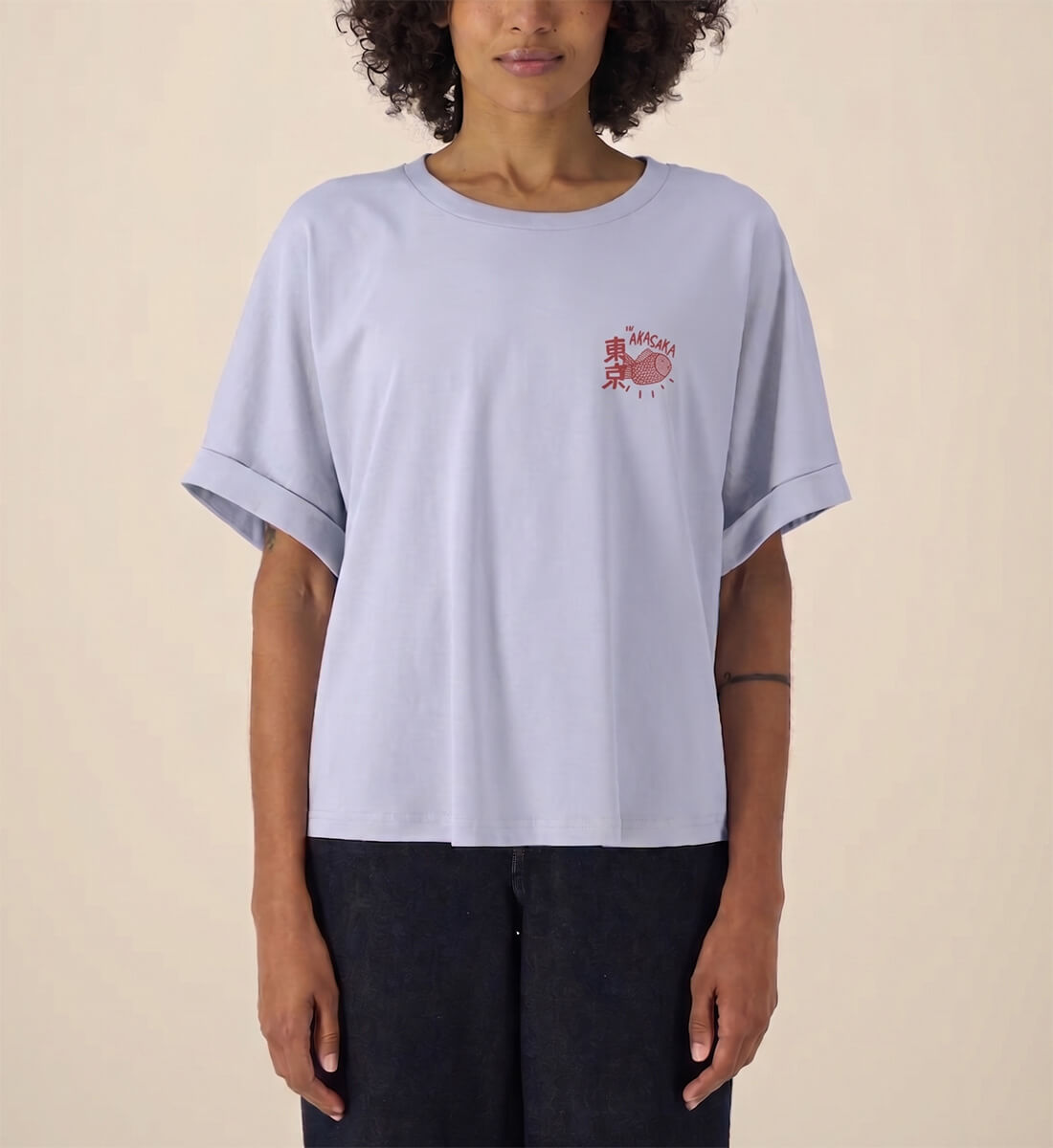 Frau trägt ein hellblaues, lässig geschnittenes Damen T-Shirt mit hochgekrempelten Ärmeln. Auf der linken Brust befindet sich ein kleiner, roter Print mit einem japanischen Taiyaki-Fisch und Kanji-Zeichen.